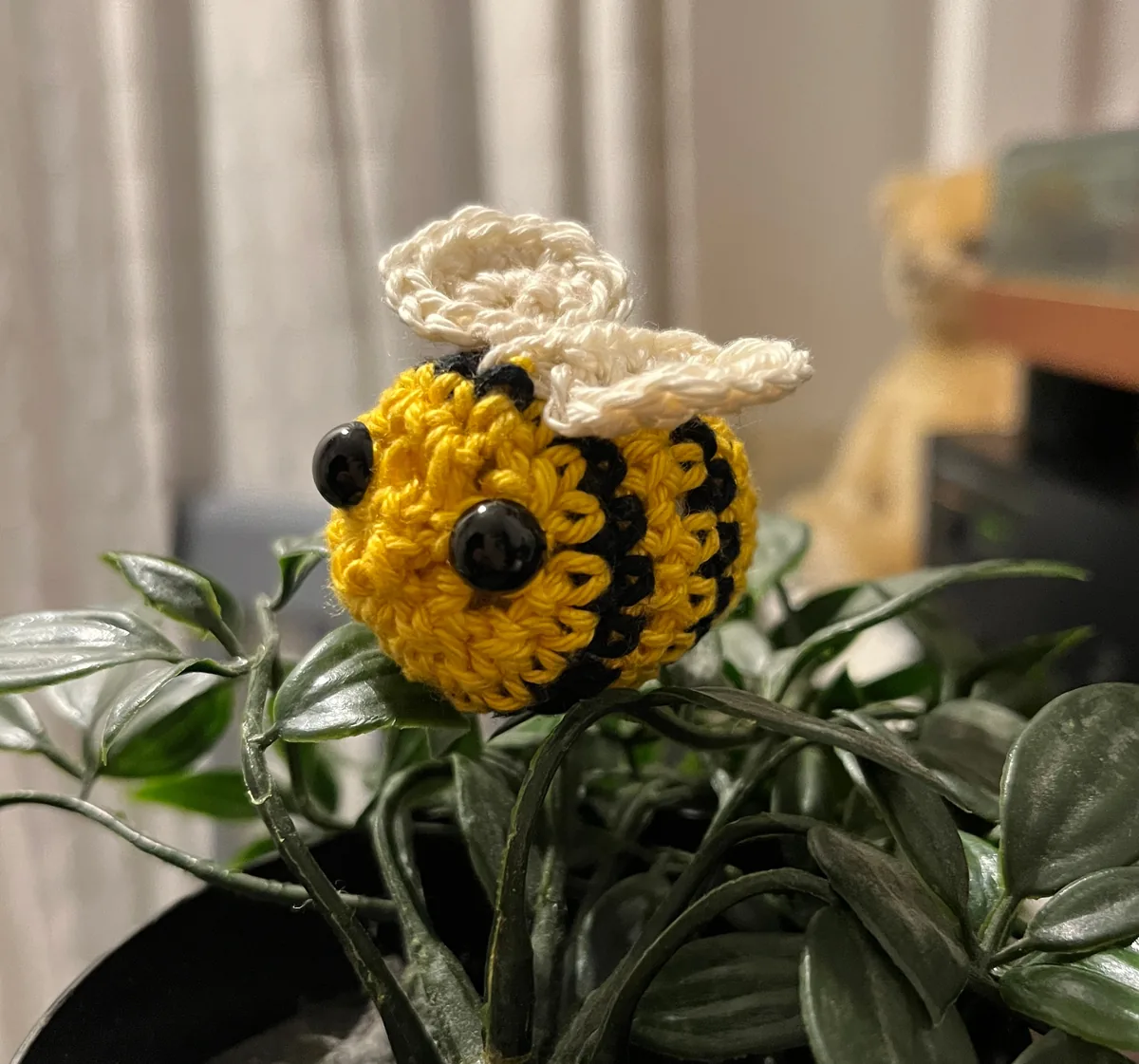 DichtBij: een klein gehaakt bijtje van gele en zwarte wol, met witte vleugels en zwarte kraaloogjes, tussen de bladeren van een plant.