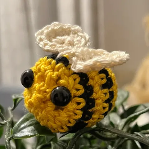 Een gehaakt bijtje van gele en zwarte wol, met witte vleugels en zwarte kraaloogjes, tussen de bladeren van een plant.