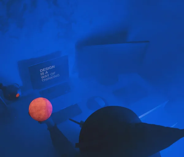 Iemand met een grote hoed houdt een oranje lichtbol vast in een blauw verlichte werkkamer
