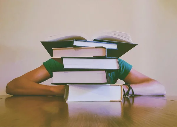 Iemand met het hoofd op tafel achter een hoge stapel boeken